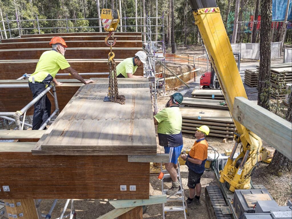 Workers guiding a large laminated veneer lumber panel lifted by crane during construction at Guulabaa.
