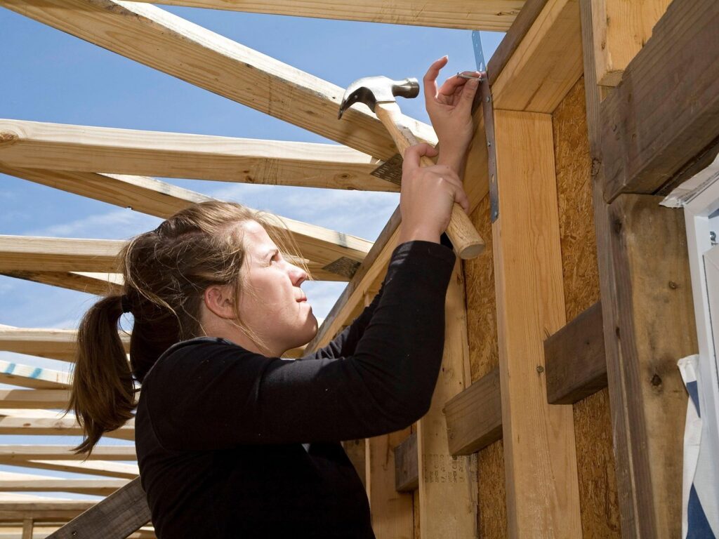 A woman hammers a metal bracket onto a timber-framed wall on a residential construction site under a blue sky