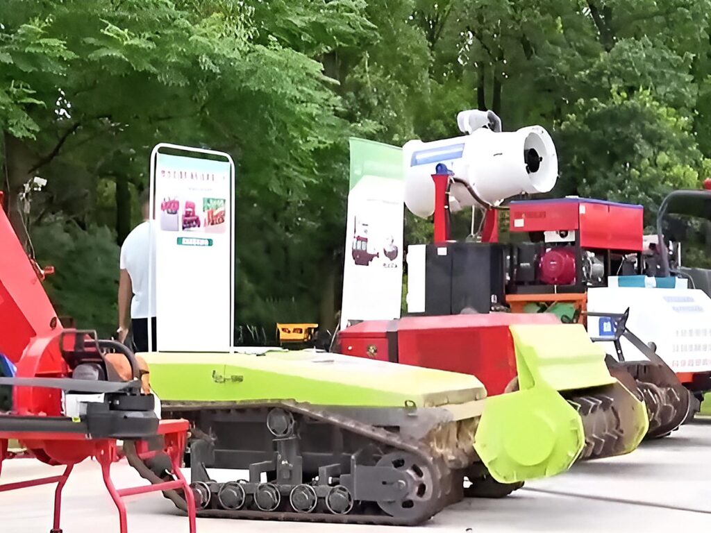 Row of red and green forestry machines including a rubber-tracked crawler and a mounted white mist blower under trees at an outdoor equipment exhibition in China.