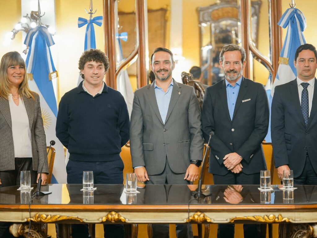 Five people stand behind a polished table in a formal room hung with Argentine flags at a government announcement