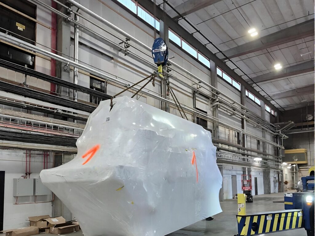 A large machine module wrapped in white plastic hangs from a crane hook inside a bright industrial hall